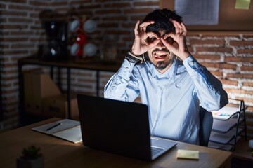 Young hispanic man with beard working at the office at night doing ok gesture like binoculars sticking tongue out, eyes looking through fingers. crazy expression.