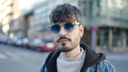 Young hispanic man standing with serious expression wearing sunglasses at street