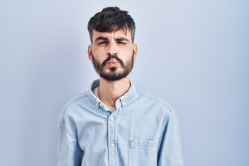 Young hispanic man with beard standing over blue background looking at the camera blowing a kiss on air being lovely and sexy. love expression.