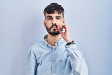 Young hispanic man with beard standing over blue background mouth and lips shut as zip with fingers. secret and silent, taboo talking