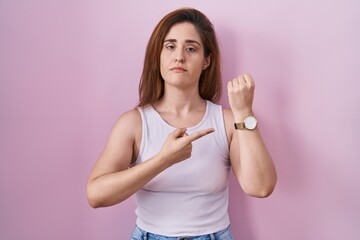 Brunette woman standing over pink background in hurry pointing to watch time, impatience, looking at the camera with relaxed expression