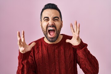 Young hispanic man with beard wearing casual sweater over pink background crazy and mad shouting and yelling with aggressive expression and arms raised. frustration concept.