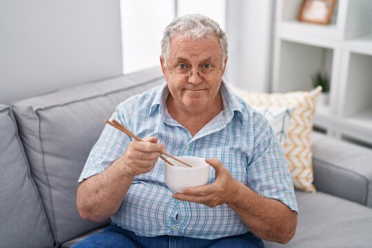 Middle age grey-haired man eating chinese food sitting on sofa at home