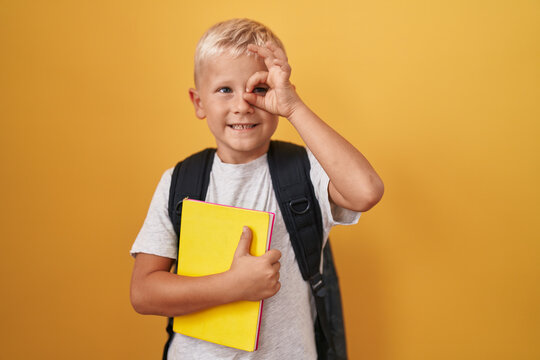 Little caucasian boy wearing student backpack and holding book smiling happy doing ok sign with hand on eye looking through fingers