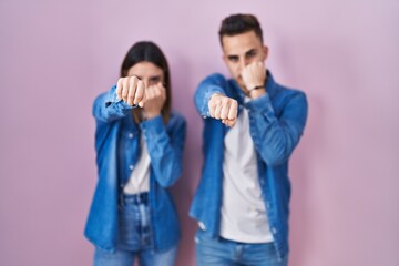 Young hispanic couple standing over pink background punching fist to fight, aggressive and angry...