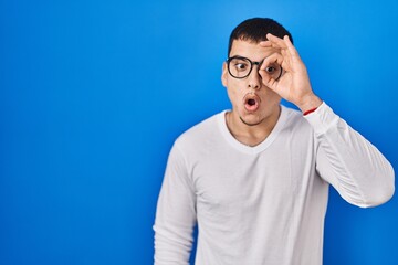 Young arab man wearing casual white shirt and glasses doing ok gesture shocked with surprised face, eye looking through fingers. unbelieving expression.