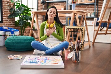 Hispanic woman sitting at art studio painting on canvas smiling with hands on chest with closed eyes and grateful gesture on face. health concept.