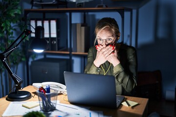 Young blonde woman working at the office at night shocked covering mouth with hands for mistake. secret concept.