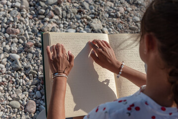 Teenage girl using both hands to read  braille book on the beach. Concept: summer reading