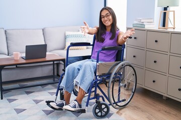 Fototapeta na wymiar Young hispanic woman sitting on wheelchair at home looking at the camera smiling with open arms for hug. cheerful expression embracing happiness.