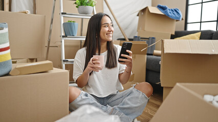 Young beautiful hispanic woman using smartphone drinking coffee at new home