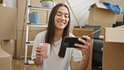 Young beautiful hispanic woman watching video on smartphone drinking coffee at new home