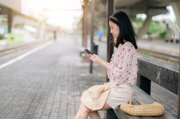 young asian woman traveler with weaving basket using a mobile phone and waiting for train in train station. Journey trip lifestyle, world travel explorer or Asia summer tourism concept.