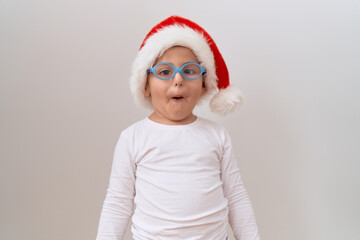 Little hispanic boy wearing glasses and christmas hat scared and amazed with open mouth for surprise, disbelief face