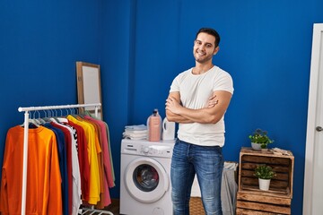 Young hispanic man standing with arms crossed gesture at laundry room