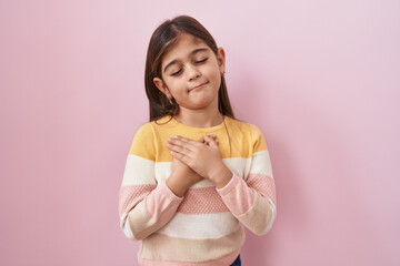 Little hispanic girl wearing sweater over pink background smiling with hands on chest, eyes closed with grateful gesture on face. health concept.