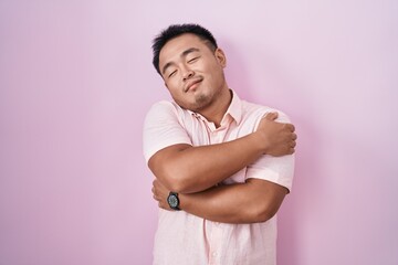 Chinese young man standing over pink background hugging oneself happy and positive, smiling...