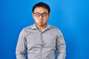 Young chinese man standing over blue background puffing cheeks with funny face. mouth inflated with...
