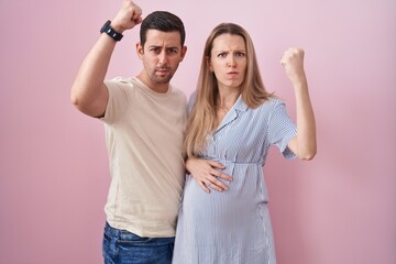 Young couple expecting a baby standing over pink background angry and mad raising fist frustrated...