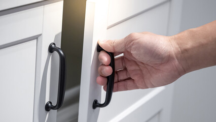 Male hand holding black door handle opening white wooden cabinet. Home furniture detail.
