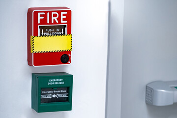 Red fire alarm switch with green box of emergency door release installed on the wall near the fire exit door. Building safety and security system