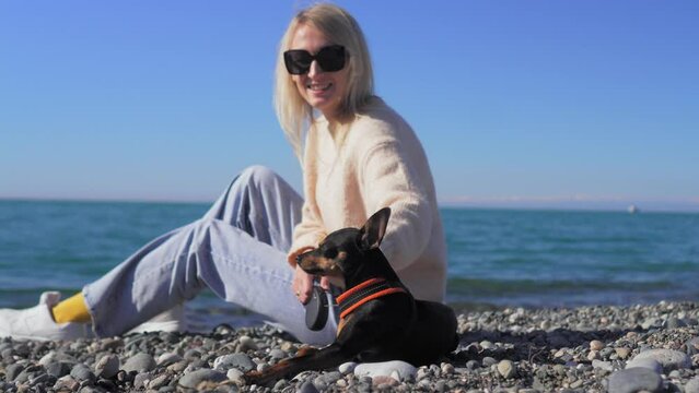 Young woman sitting on beach with her little dog Toy Terrier. Concept friendship - woman and dog sit together on beach and enjoy weekend and wonderful day against backdrop sea or ocean. vertical video