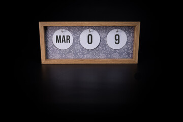 A wooden calendar block showing the date March 9th on a dark black background, save the date or...
