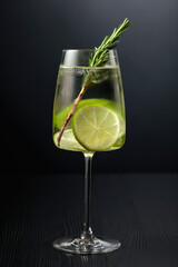 Gin tonic with ice, rosemary, and lime in frosted glass on a black table.