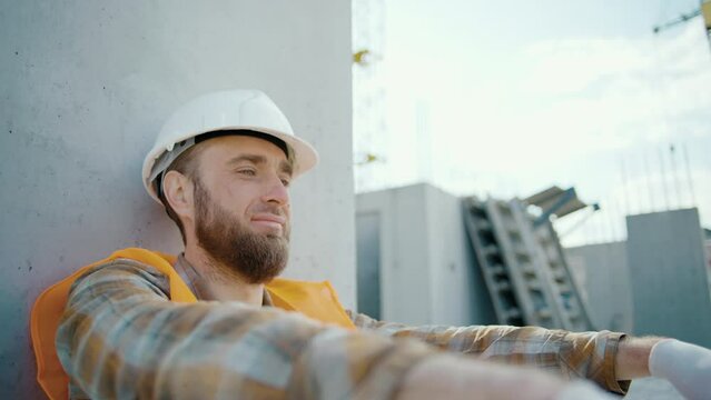 Young foreman sitting outdoors and feeling tired after overwork. Caucasian worker in reflective vest and helmet at construction site. Slow motion
