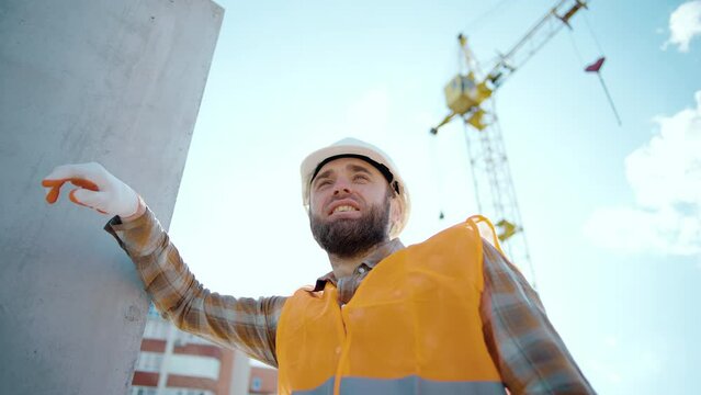 A tired worker at a construction site rests his hand on the wall after hard work and is under stress. Worker during a break at the construction of a road on a sunny summer day. Slow motion