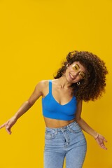 Woman with curly afro hair in a blue T-shirt on a yellow background dancing flying hair with sunglasses yellow, hand signs, look into the camera, smile with teeth and happiness, copy space