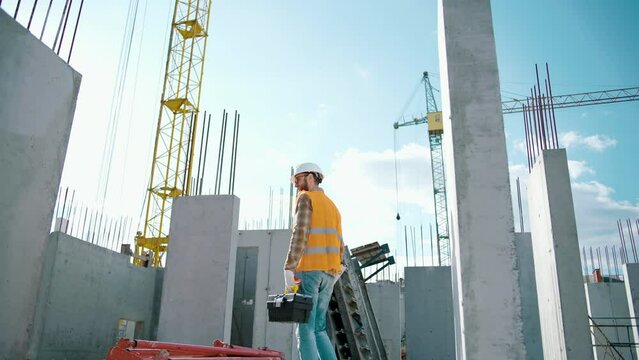 Construction worker walking and working on a construction site with a toolbox and bubble level and high visibility clothing. Man turns to the camera