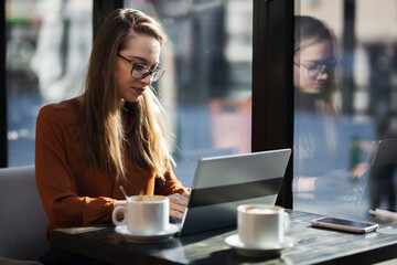 Young businesswoman in a cafe bar or restaurant. Freelancer girl working on laptop and having tea at a window table.