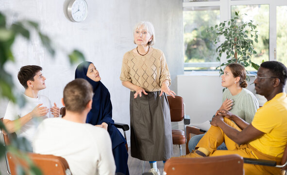 Friendly and calm senior female psychotherapist or facilitator leading group therapy session with women and men of different ages  - Powered by Adobe
