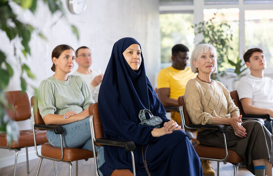 Focused young asian girl dressed in traditional muslim abaya and chador sitting in lecture hall with group of men and women 