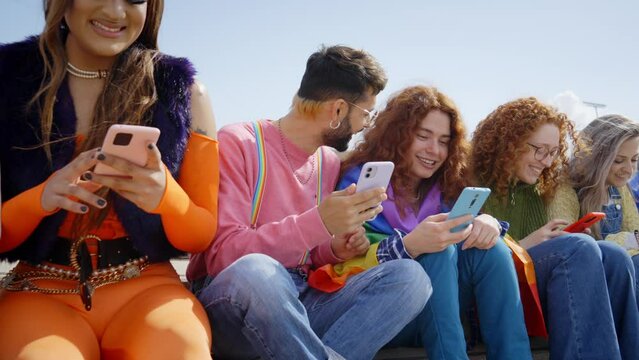 Group of young friends from LGBT community using mobile phone and celebrating gay pride festival day together. Happy people gathered sitting outdoors checking social networks. Generation z and apps.