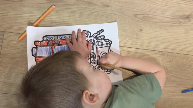 toddler baby child drawing coloring fire truck lying on wooden floor at home. male kid 4 years tired lie down. little boy in green t-shirt doing homework art class. hardworking child. hobby education 