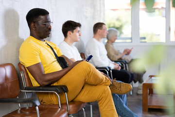Adult African American in casual clothes sitting in lobby of medical clinic, browsing Internet while waiting for appointment with doctor