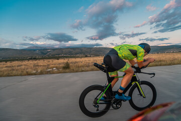 Naklejka premium Triathlete riding his bicycle during sunset, preparing for a marathon. The warm colors of the sky provide a beautiful backdrop for his determined and focused effort.
