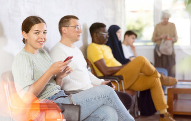 Casual positive young girl sitting in lobby of medical clinic, browsing web pages in Internet on...