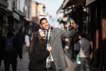 Multiracial couple posing on narrow streets in old part of the sity. Taking selfy photo with phone....