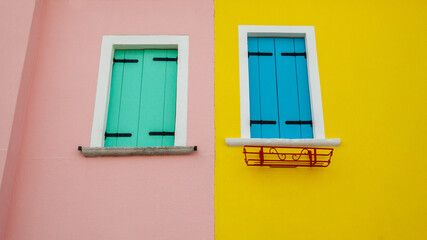 Two windows with green and blue shutters on pink and yellow walls, next door to each other