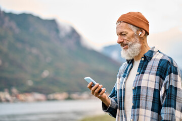 Cool bearded healthy older elder hipster man standing in nature park wearing earbud using mobile...