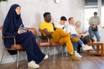 Young Asian girl in black abaya and khimar sitting in lobby of job center or employment office,...