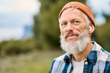 Cool bearded healthy older elder hipster man standing in fall nature park wearing earbud. Mature traveler enjoying freedom, active leisure lifestyle, listening music playlist, portrait.