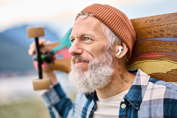 Active cool bearded old hipster man standing in nature park holding skateboard wearing earbud....