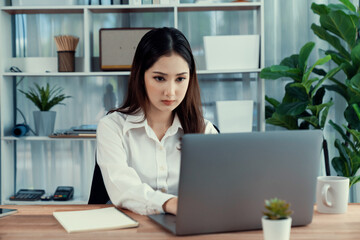 Young asian enthusiastic businesswoman at modern office desk using laptop to work with a cup of coffee. Diligent and attractive office lady working on computer notebook in her office work space.