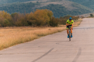 Obraz premium Triathlete riding his bicycle during sunset, preparing for a marathon. The warm colors of the sky provide a beautiful backdrop for his determined and focused effort.