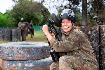 Fototapeta premium Young smiling woman paintball player in camouflage posing with gun outdoors