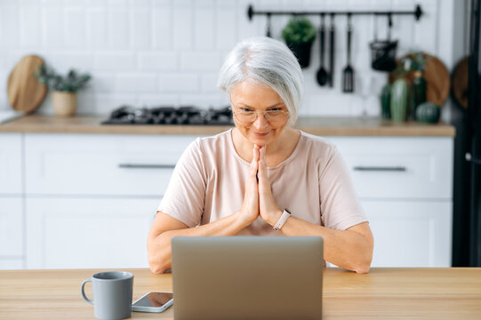 Pretty, modern positive caucasian gray haired woman with glasses, sitting at home in the kitchen at the table, looks at the laptop screen, wants something, worries, holds palms together, prays